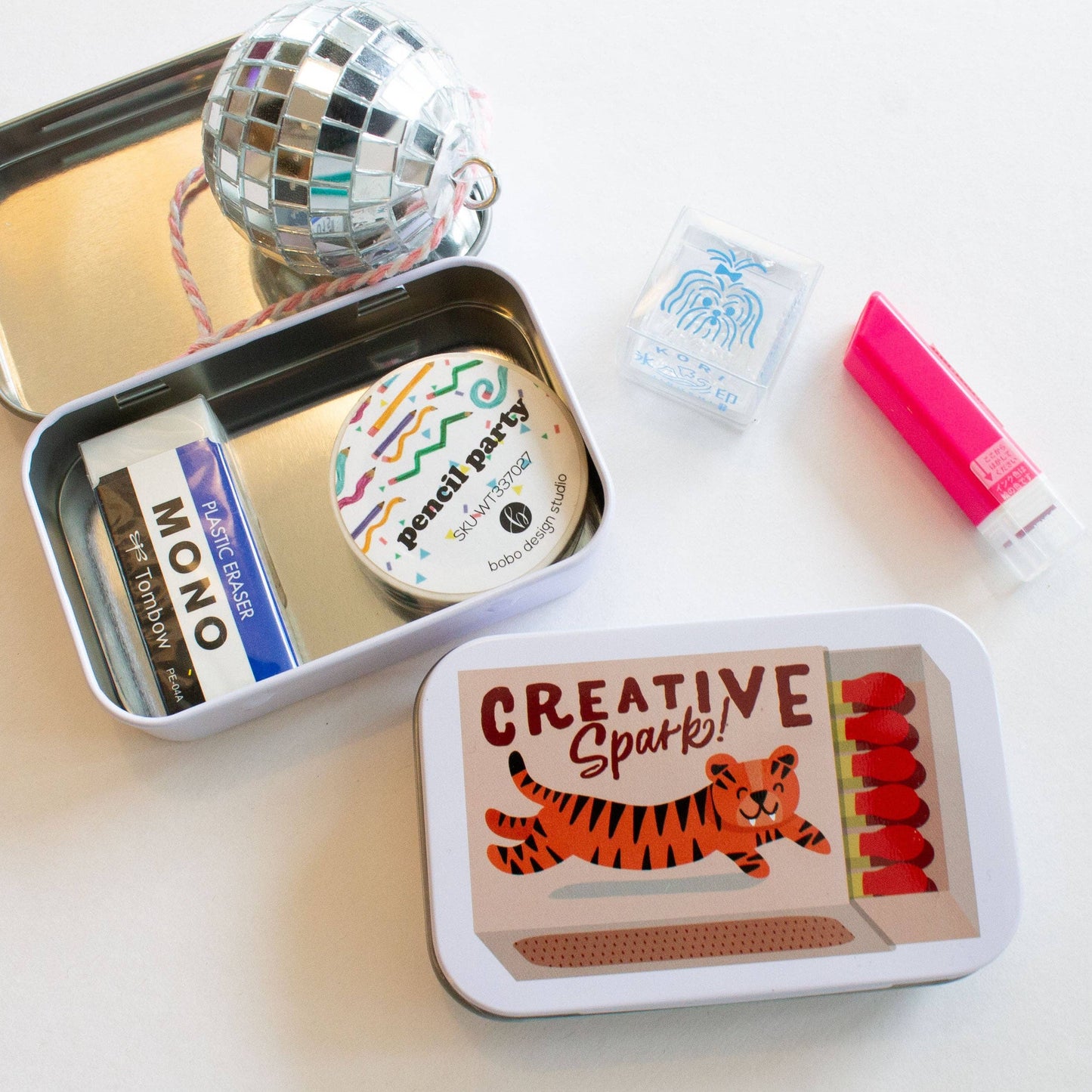 Small tin with stationery items including a eraser, marker, and small disco ball on a white surface.