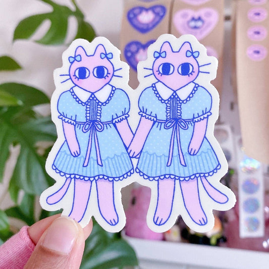 Two stickers of cartoon cat girls in blue dresses held by a hand against a blurred background. Creepy Cat Twins Sticker