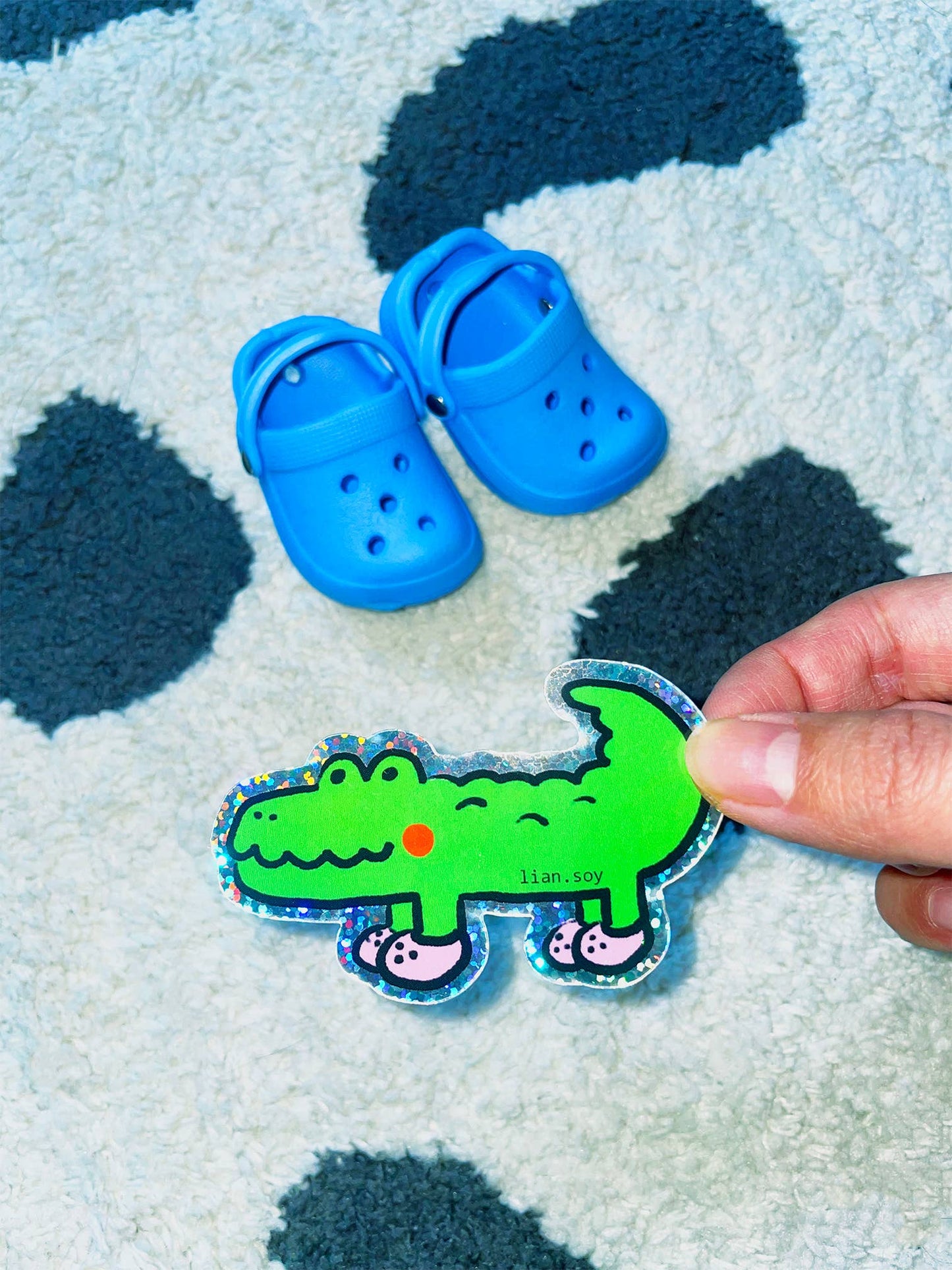 Blue children's shoes on a textured surface with a hand holding a green dinosaur sticker.