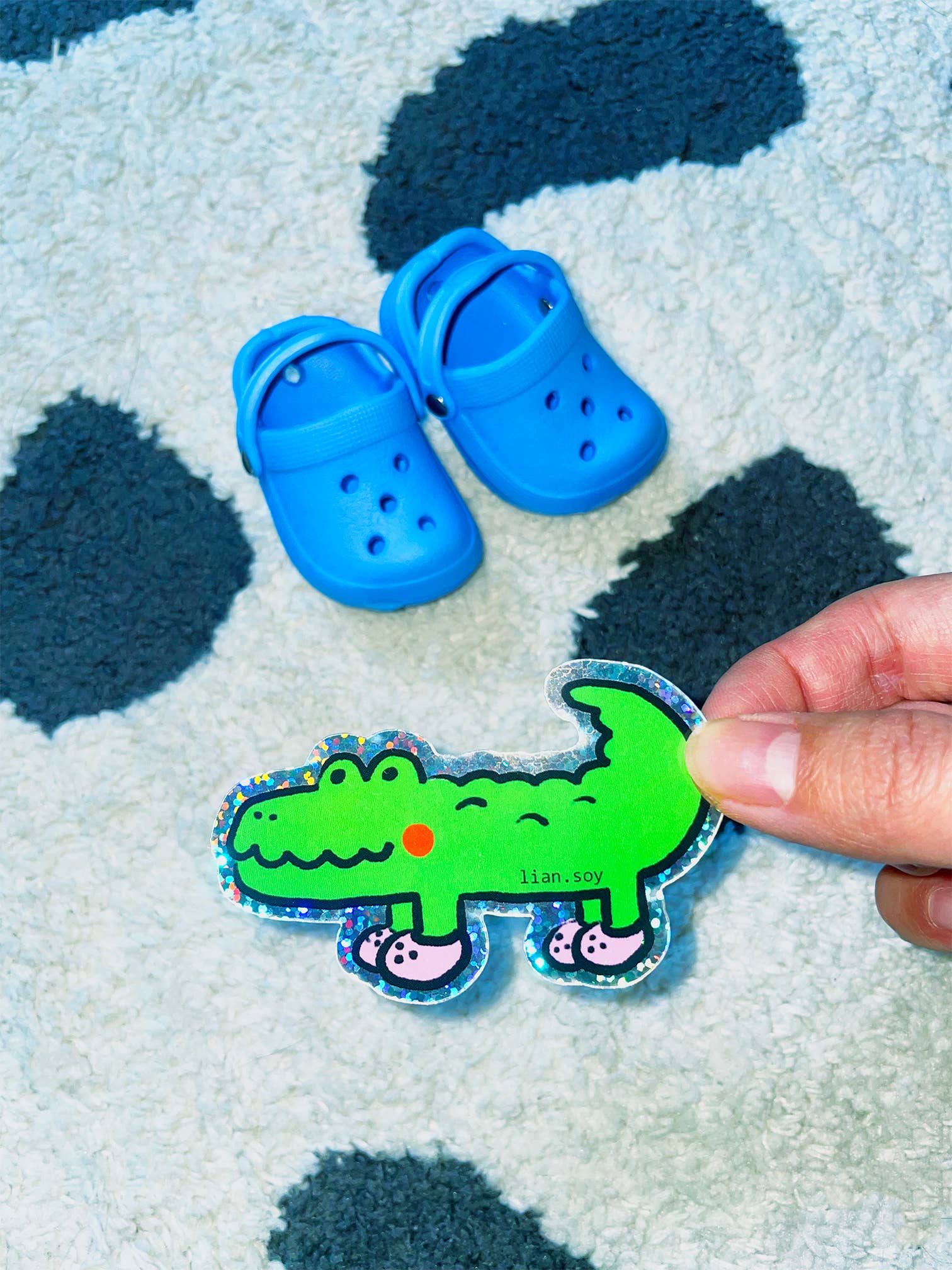 Blue children's shoes on a textured surface with a hand holding a green dinosaur sticker.