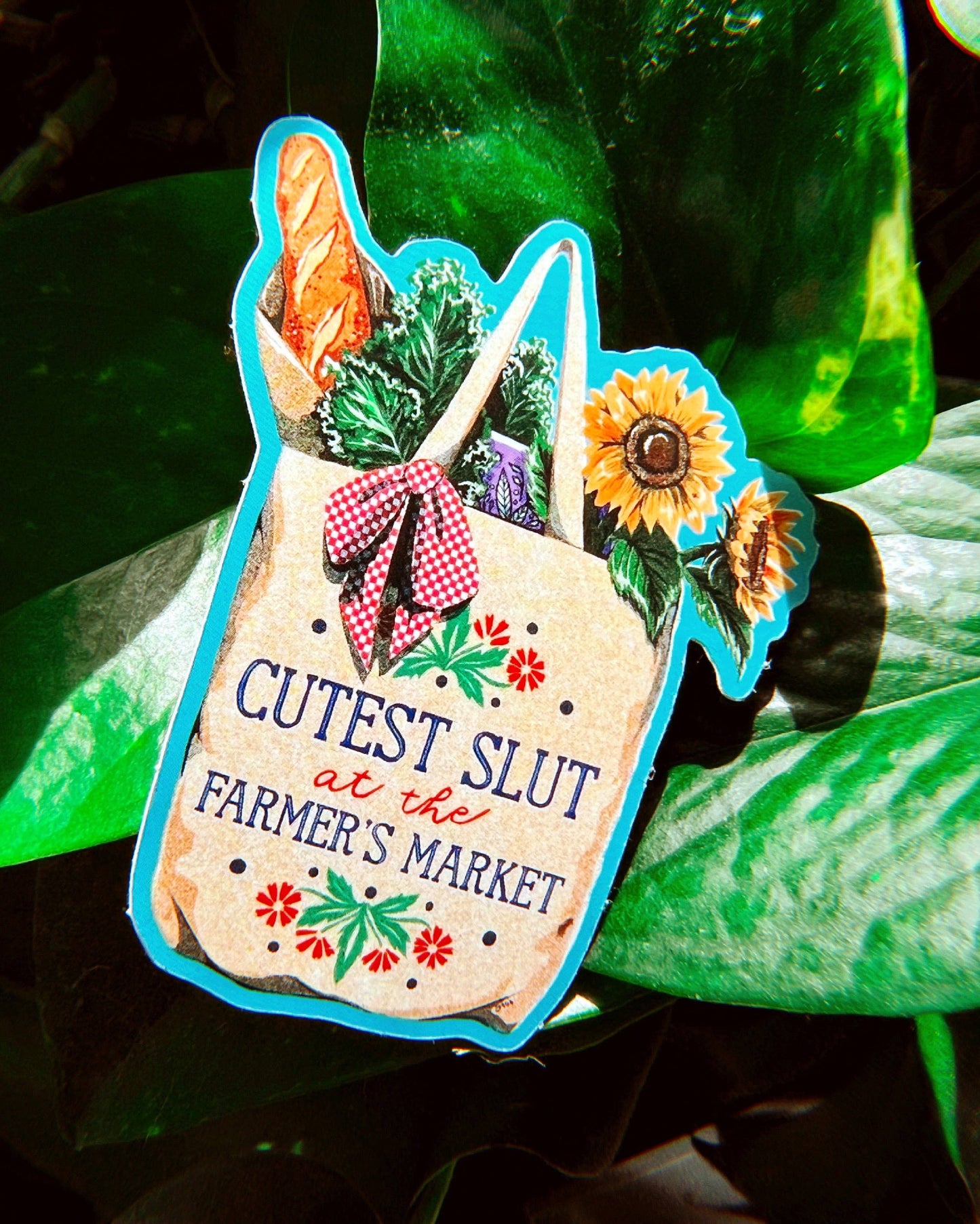 Sticker with a design of a bag of groceries and text 'Cutest Slut at the Farmer's Market' against a green leafy background.