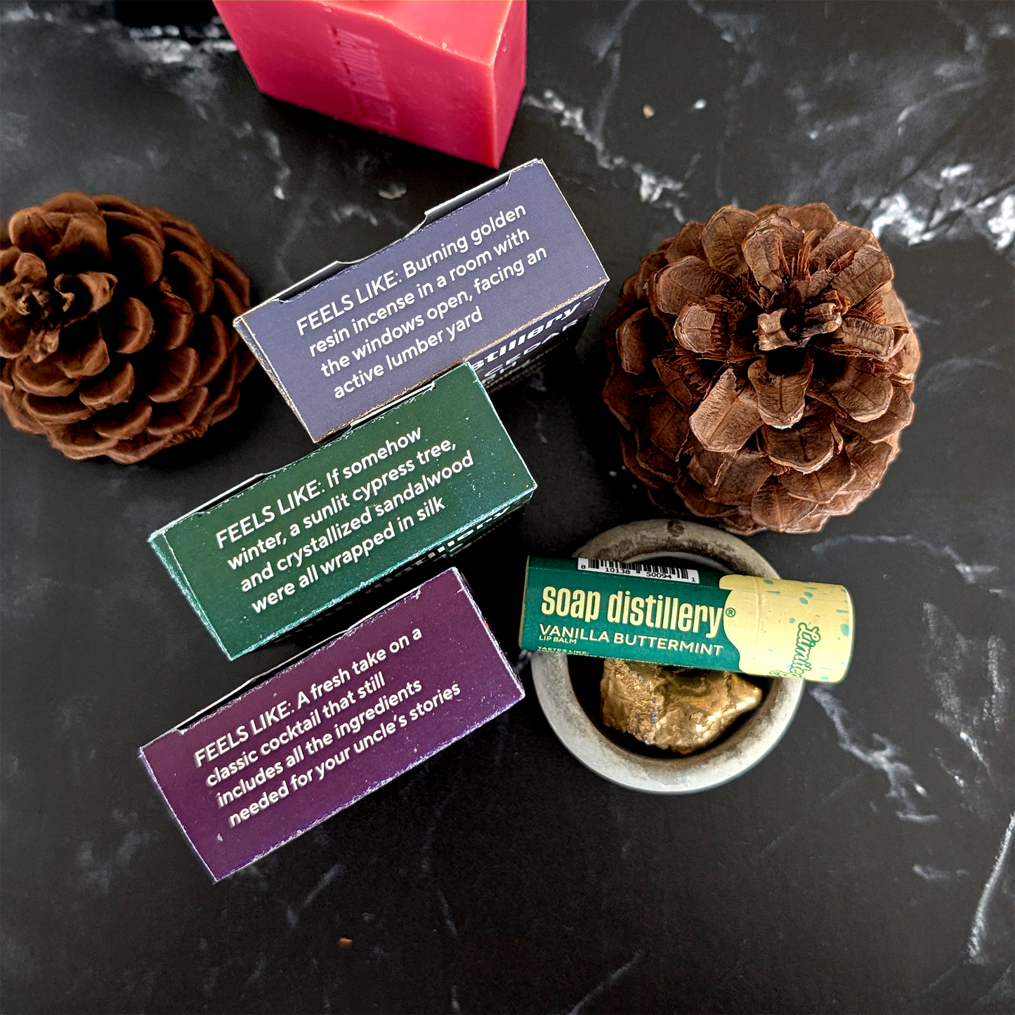A styled flat-lay of a soap and candle set on a dark marble surface, featuring three wooden boxes with colorful labels, a pink bar of soap, a small bowl of wax melt, and two pinecones.