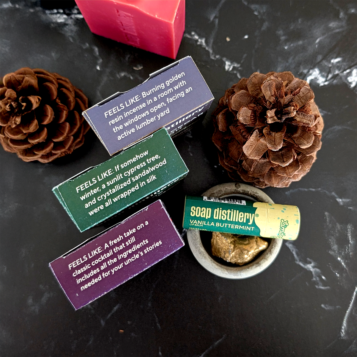 A styled flat-lay of a soap and candle set on a dark marble surface, featuring three wooden boxes with colorful labels, a pink bar of soap, a small bowl of wax melt, and two pinecones.