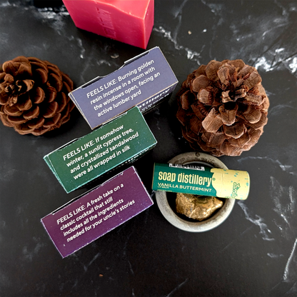 A styled flat-lay of a soap and candle set on a dark marble surface, featuring three wooden boxes with colorful labels, a pink bar of soap, a small bowl of wax melt, and two pinecones.