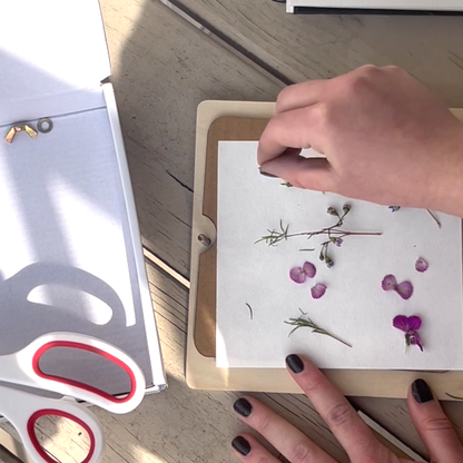 Person pressing flowers onto a card with a pair of scissors nearby on a wooden surface