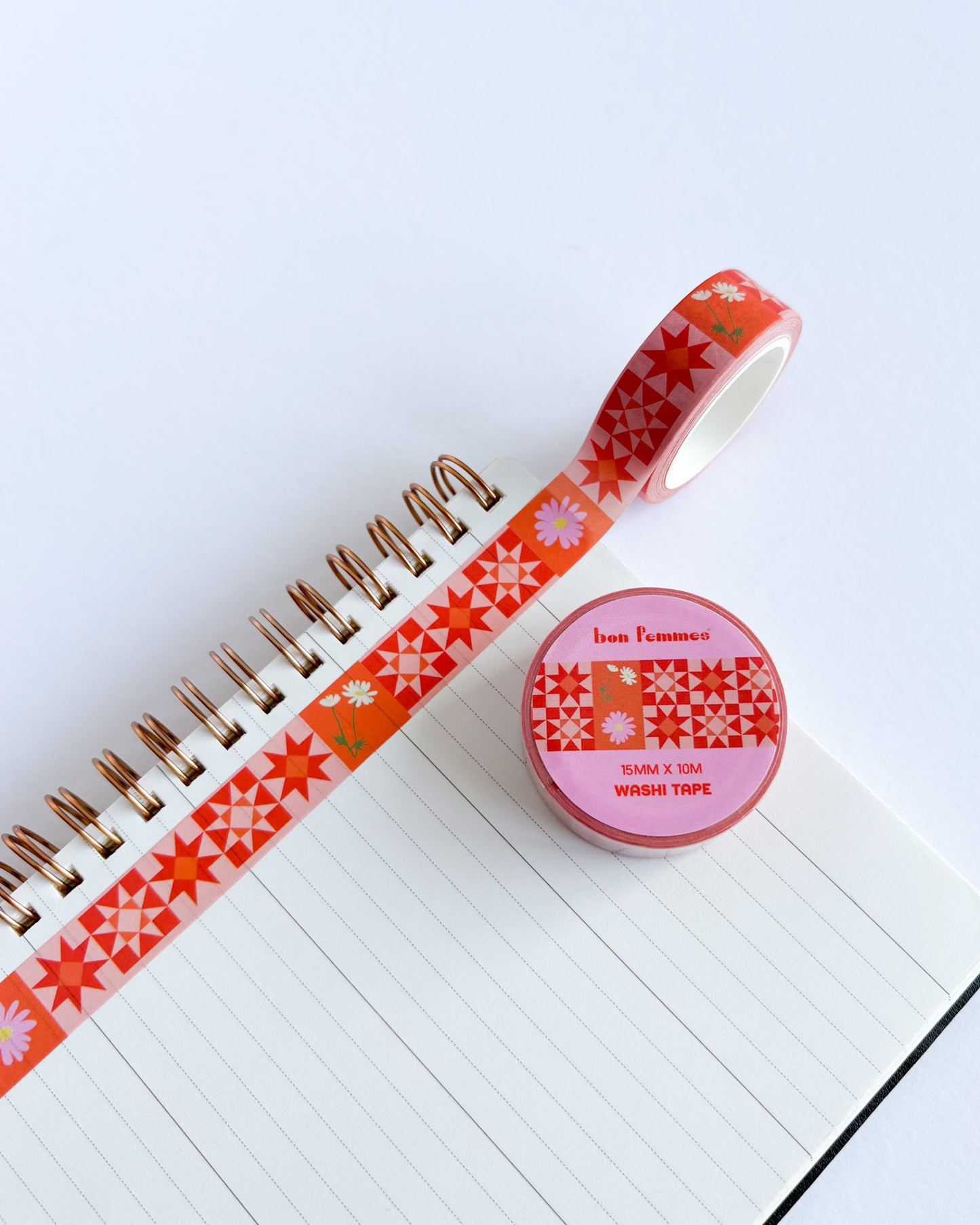 A roll of decorative washi tape with a red and orange geometric pattern is partially unrolled across an open lined notebook. The matching roll of tape sits on the notebook cover, showing the brand label 'bon l'homme'.