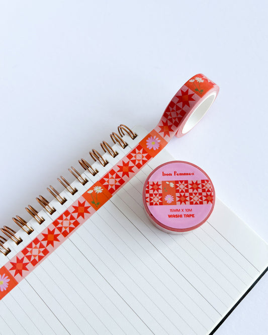 A roll of decorative washi tape with a red and orange geometric pattern is partially unrolled across an open lined notebook. The matching roll of tape sits on the notebook cover, showing the brand label 'bon l'homme'.