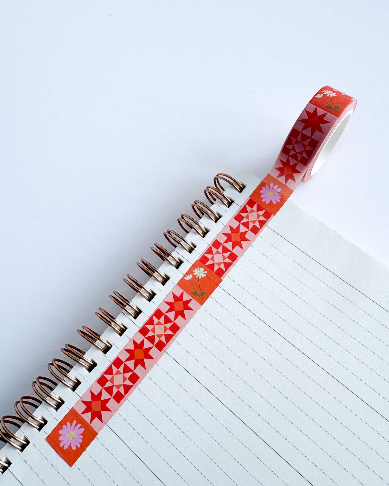 A roll of decorative washi tape with a red and orange geometric star pattern and small white flowers, shown partially unrolled over an open spiral notebook on a white background.