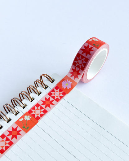 A roll of decorative washi tape with a red and orange geometric star pattern and small daisy illustrations, shown partially unrolled next to a spiral notebook on a light blue background.