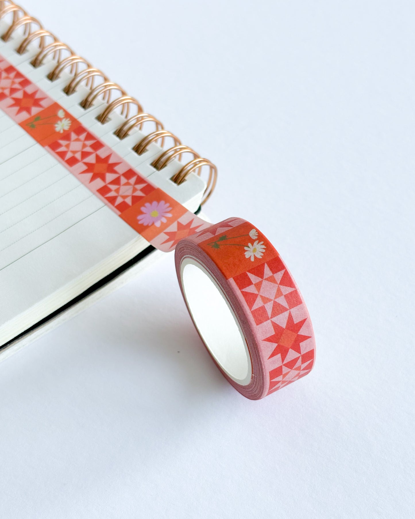A roll of decorative washi tape with a red and pink geometric star pattern and small white flowers, shown partially unrolled across an open spiral notebook on a clean white background.