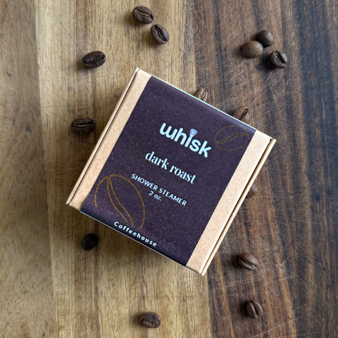 A square cardboard box of Coffeehouse Whisk Dark Roast Shower Steamer, placed on a rustic wooden surface with scattered coffee beans around it.