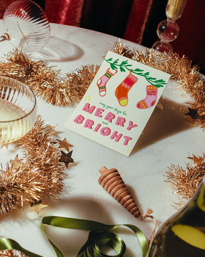 Christmas card with colorful socks and 'Merry Bright' text on a table with festive decorations. Days be Merry & Bright Holiday Greeting Card
