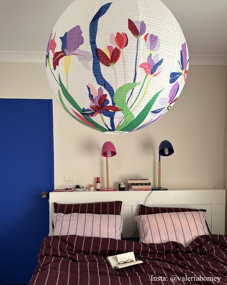 A large white paper lantern-style pendant light with a colorful floral print hangs in a bedroom above a bed with burgundy striped bedding. The room has beige walls, a blue door, and two small table lamps on a shelf.