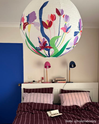 A large white paper lantern-style pendant light with a colorful floral print hangs in a bedroom above a bed with burgundy striped bedding. The room has beige walls, a blue door, and two small table lamps on a shelf.