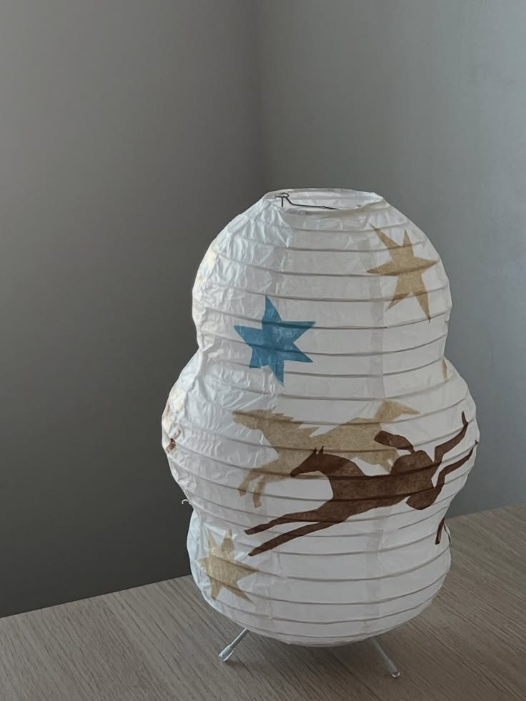 A white paper lantern with a ribbed texture, decorated with hand-painted stars and a brown reindeer silhouette, displayed on a light wood surface against a gray wall.