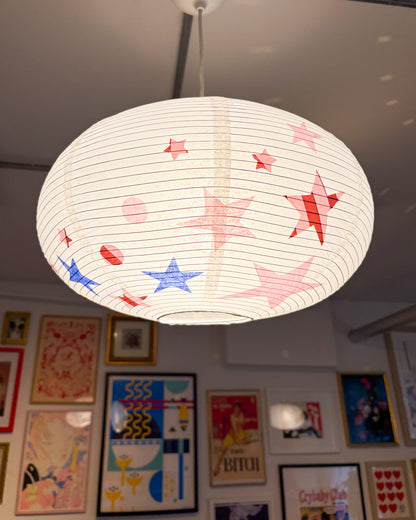 A round white paper lantern-style pendant light with red and blue star and dot patterns, hanging in a room with framed art on the wall.