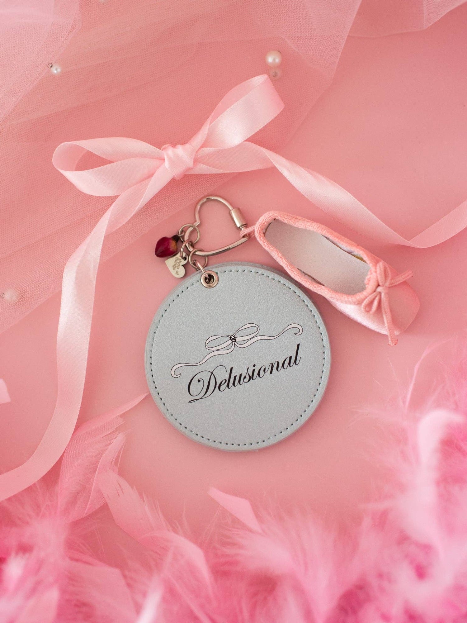 Round keychain with 'Delusional' branding on a pink background with ribbons and feathers.