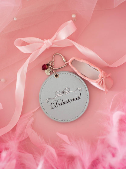 Round keychain with 'Delusional' branding on a pink background with ribbons and feathers.