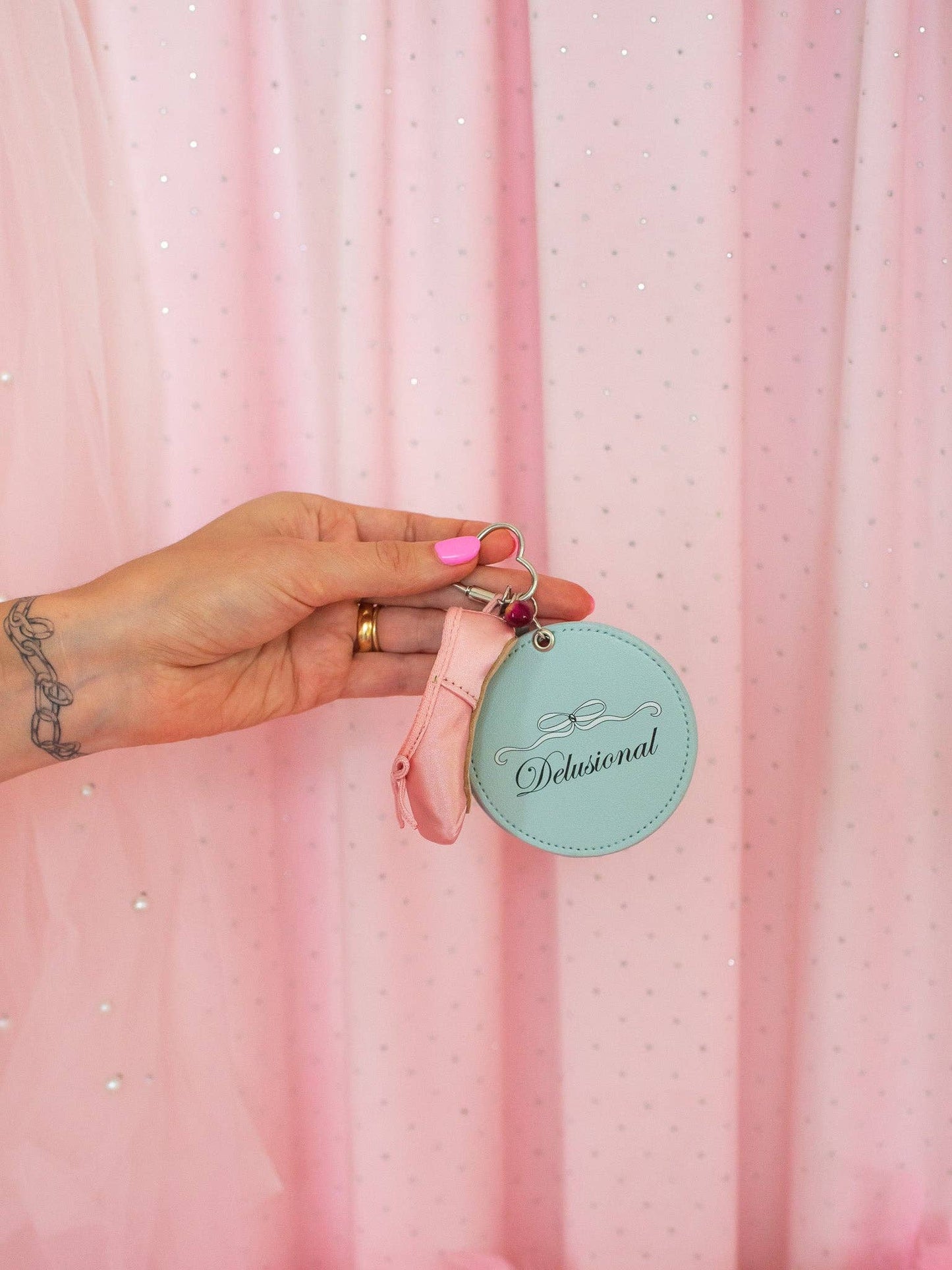 Hand holding a pink keychain with a teal tag against a pink curtain background Delusional Mirror Keychain