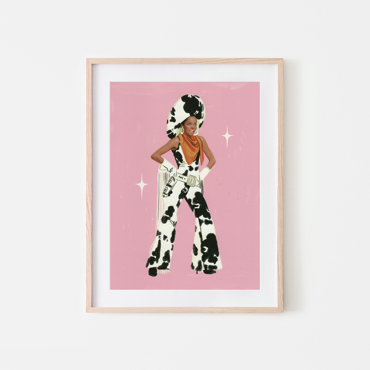 A framed art print featuring a stylized illustration ofDIANA ROSS in a black-and-white cow-print outfit against a pink background, displayed in a light wood frame on a white wall.
