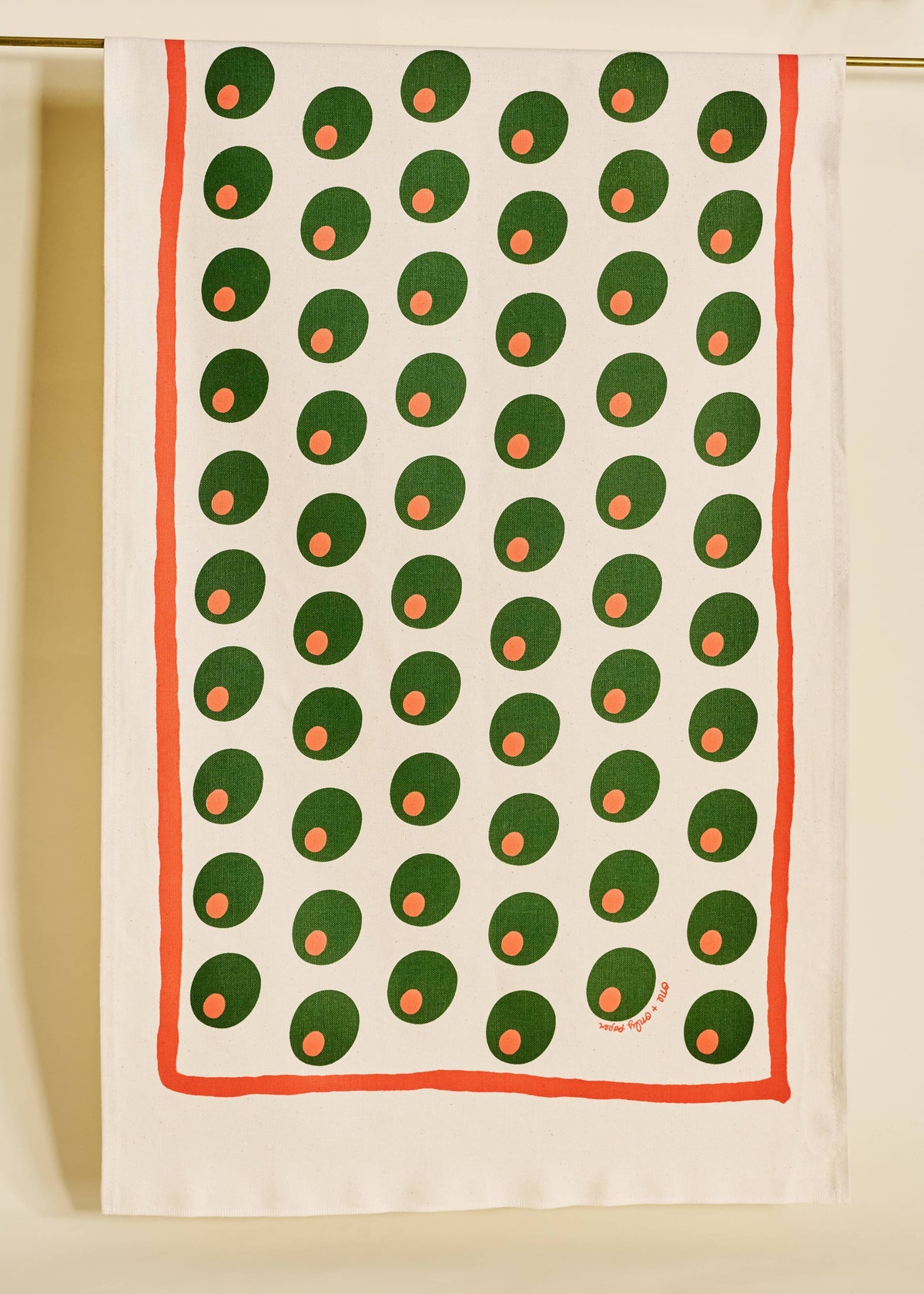 An off-white tea towel with a repeating pattern of green circles containing orange dots, framed by a red-orange border, hanging against a neutral beige background.