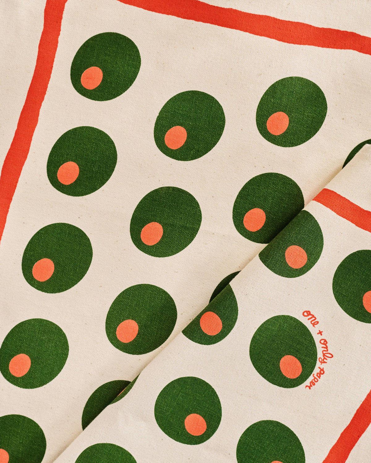 A close-up of a scarf featuring a cream background with green circles containing small orange centers, bordered by a red-orange line, and a folded section showing the brand name 'O'N + O'NLY O'N'.