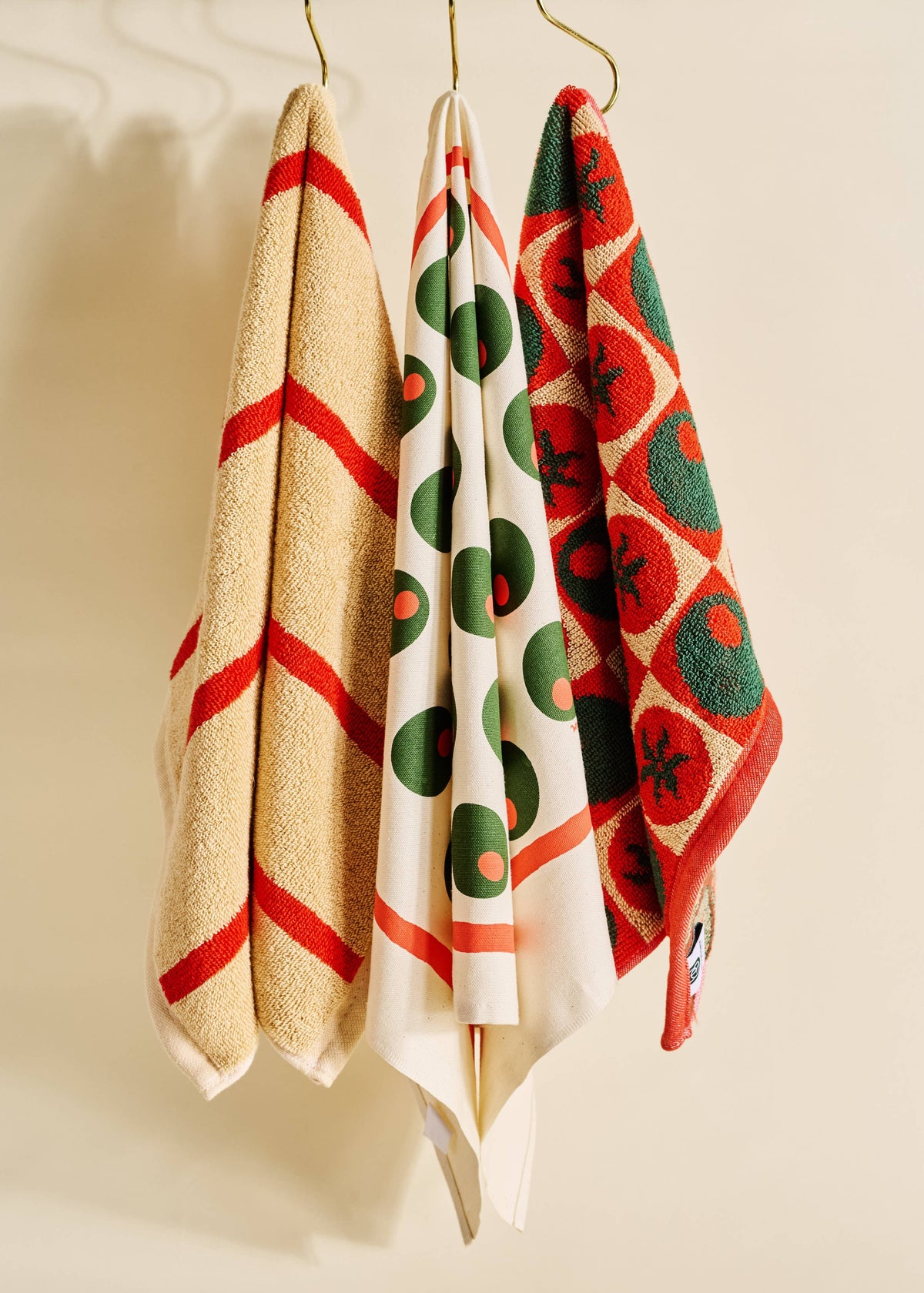 Three patterned cotton towels hanging from gold hooks against a warm cream wall. The left towel is beige with bold red diagonal stripes.