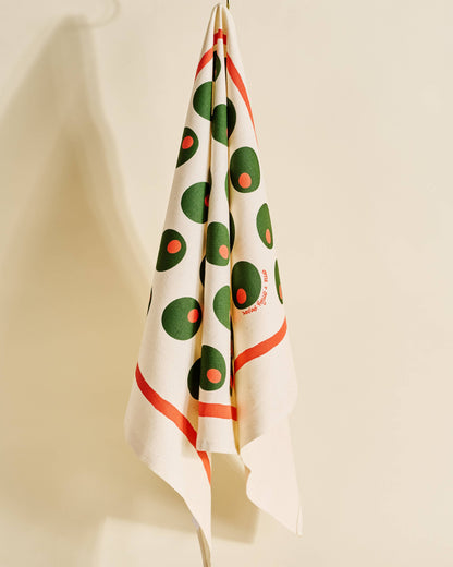 A cream-colored kitchen towel with a retro pattern of green and orange circles, hanging from a gold hook against a warm beige wall.