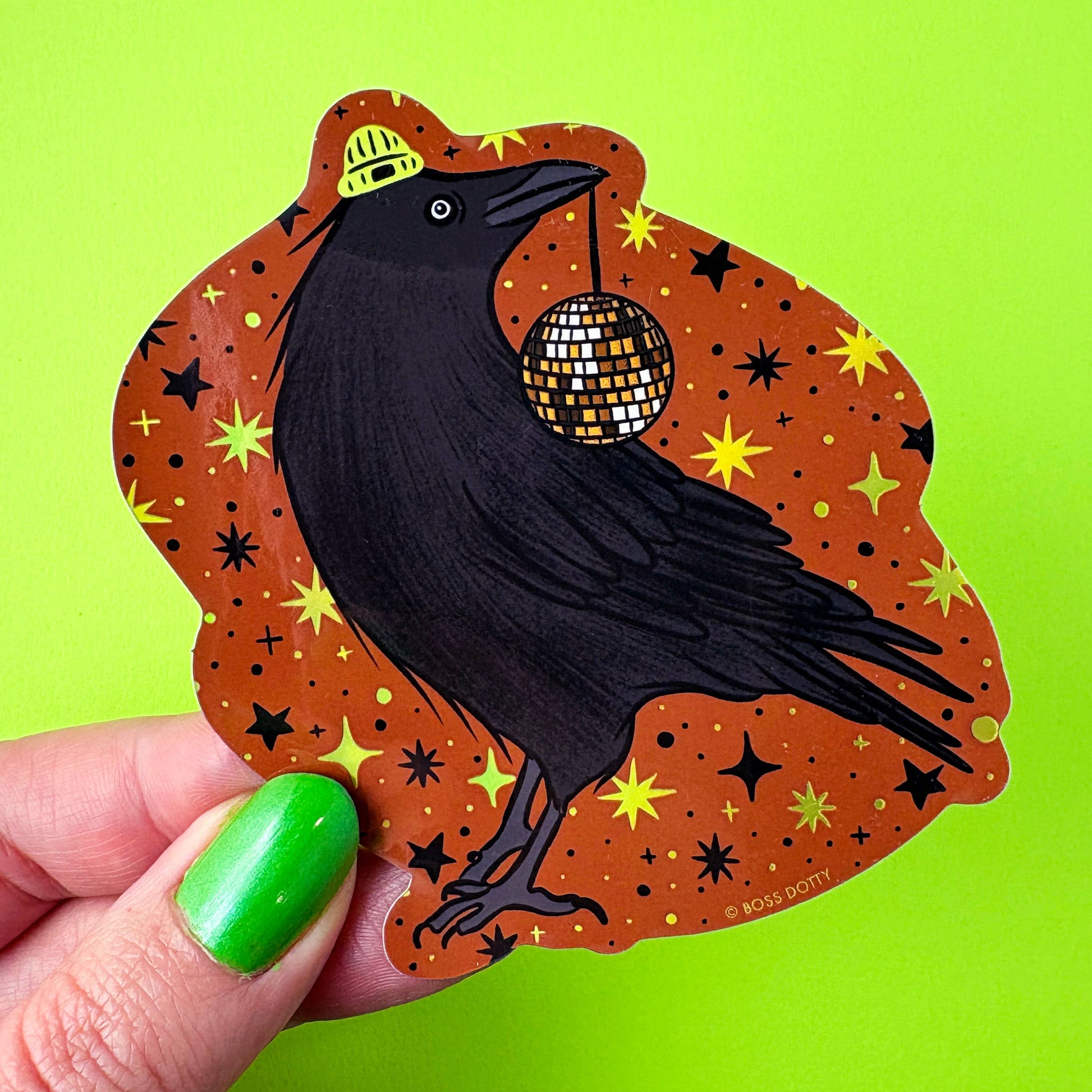Sticker of a black bird with a disco ball on an orange background held by a hand with green nail polish.