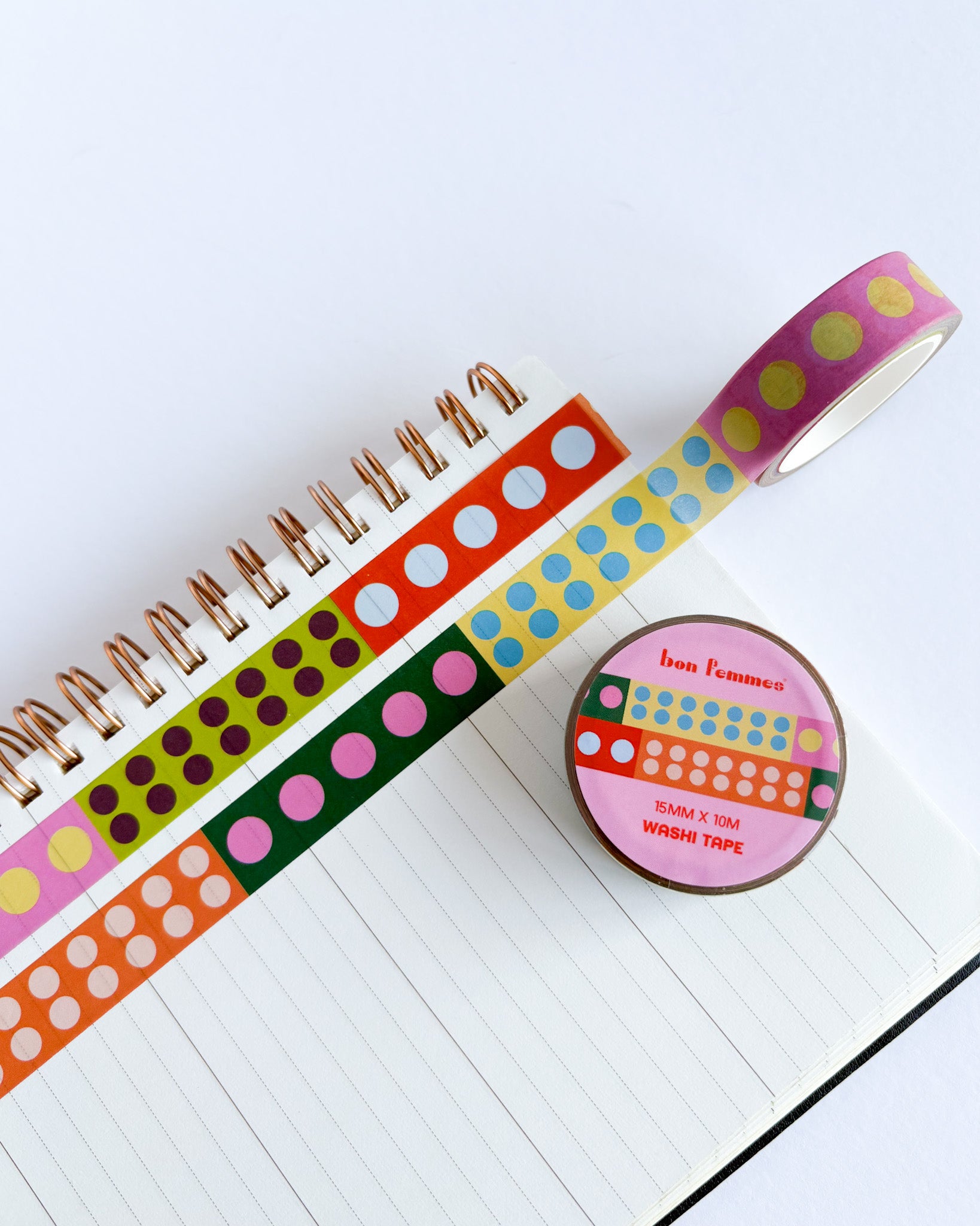 A roll of multicolor washi tape with yellow polka dots on a pink background, shown next to several other patterned washi tapes on an open lined notebook. A round label on the tape roll reads 'bean tomatoes'.