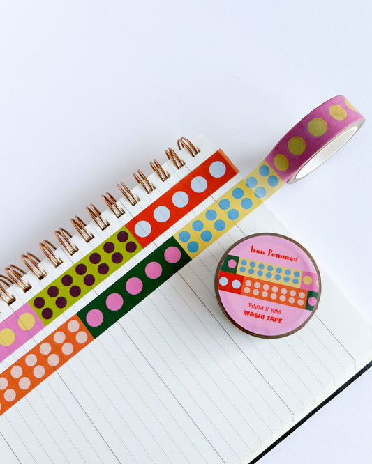 A roll of multicolor washi tape with yellow polka dots on a pink background, shown next to several other patterned washi tapes on an open lined notebook. A round label on the tape roll reads 'bean tomatoes'.
