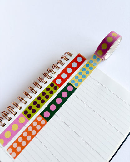 A roll of decorative washi tape with colorful polka dots is partially unrolled over an open lined notebook. The tape features multiple colors including pink, orange, green, blue, yellow, and purple, each with different dot patterns.