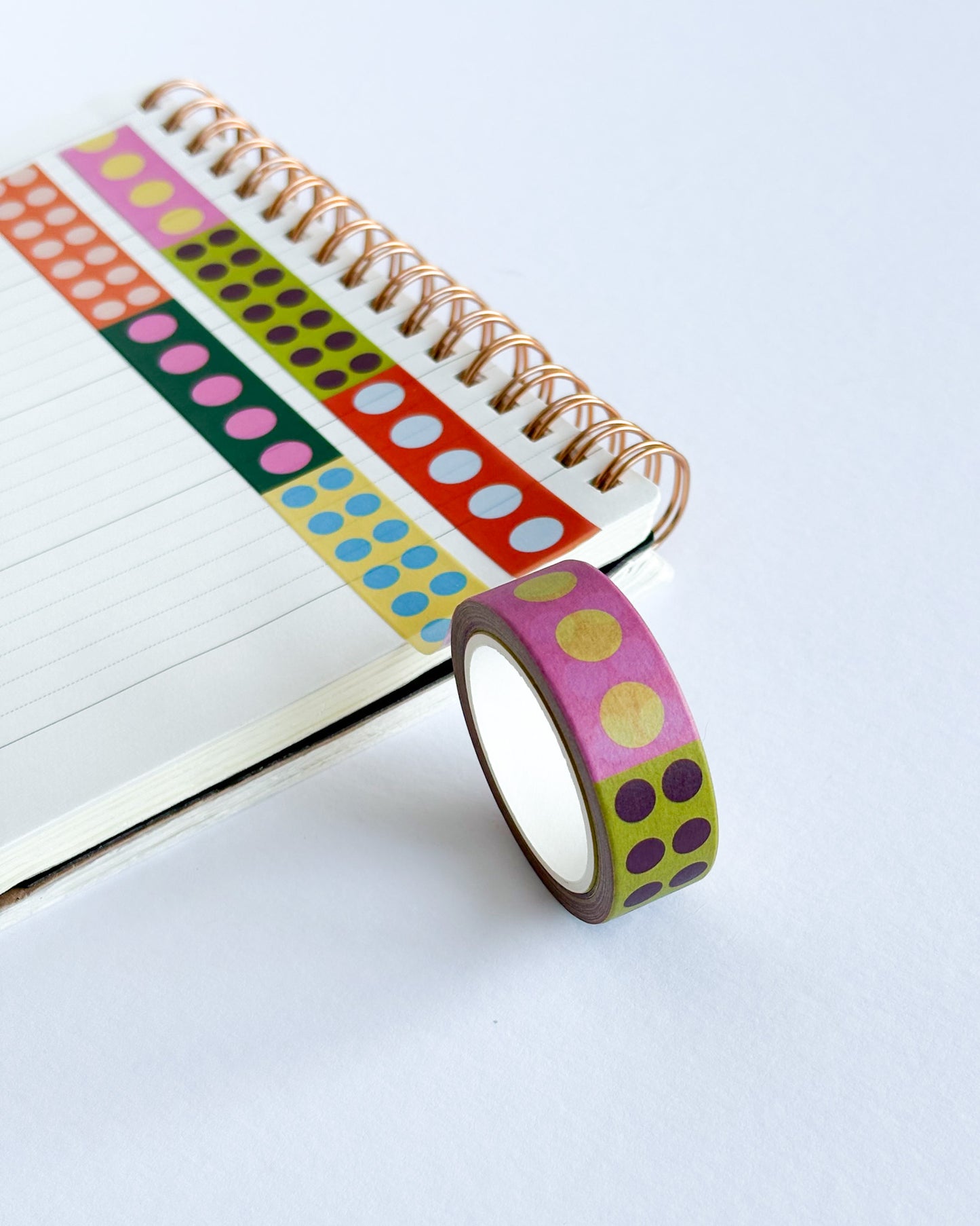A roll of decorative washi tape with pink, yellow, and purple polka dots on a lime green background, shown next to an open spiral notebook on a white surface.