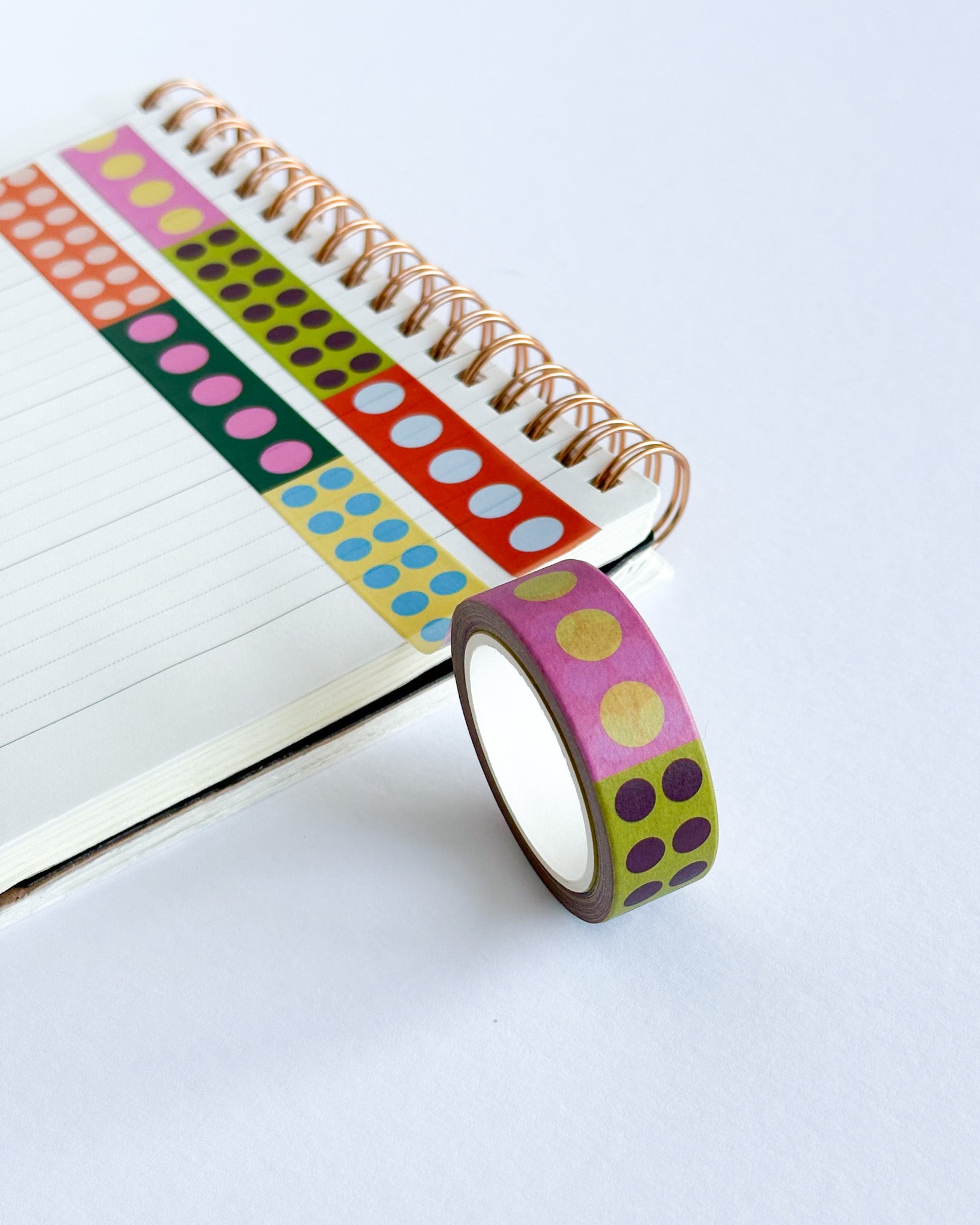 A roll of decorative washi tape with pink, yellow, and purple polka dots on a lime green background, shown next to an open spiral notebook on a white surface.