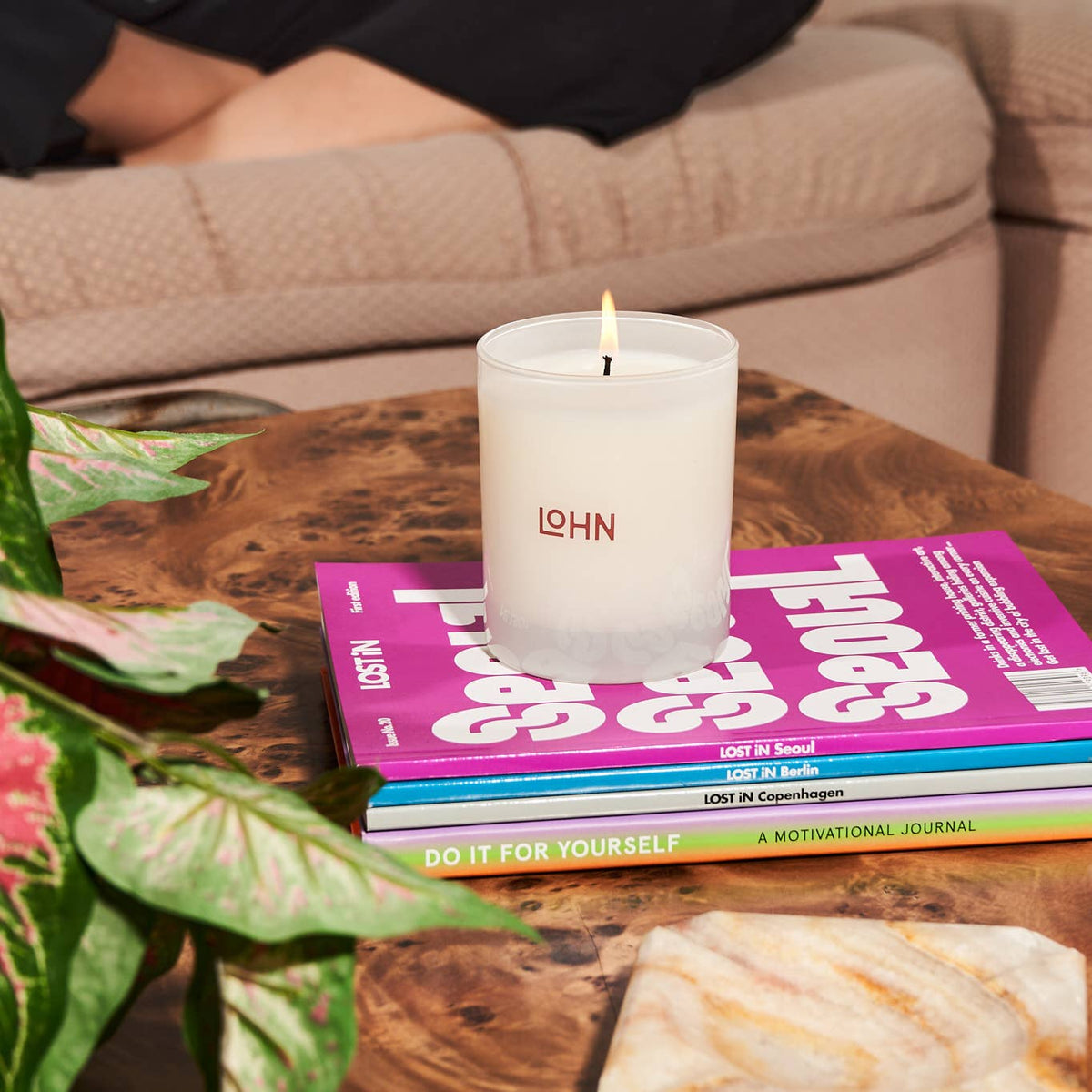 Candle with 'LOHN' branding on a stack of books on a wooden table, with a plant and marble object in the foreground.