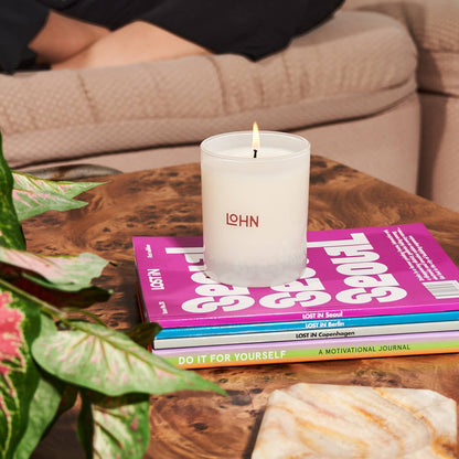 Candle with 'LOHN' branding on a stack of books on a wooden table, with a plant and marble object in the foreground.