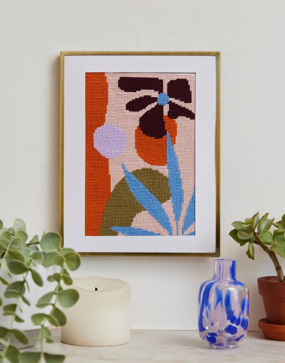 Framed cross-stitched artwork with abstract leaves on a wall with plants and a candle.