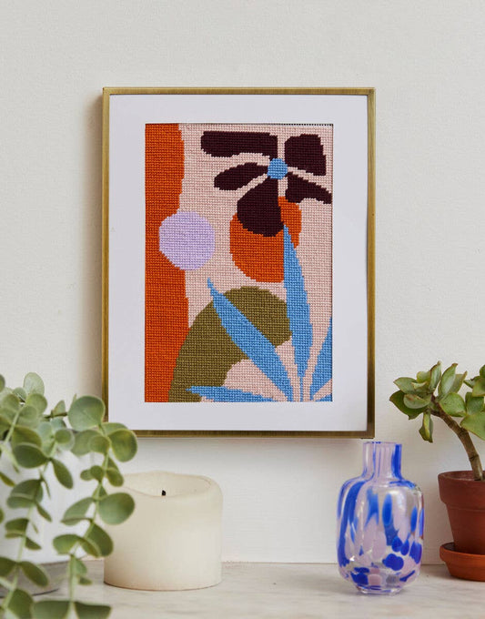 Framed cross-stitched artwork with abstract leaves on a wall with plants and a candle.