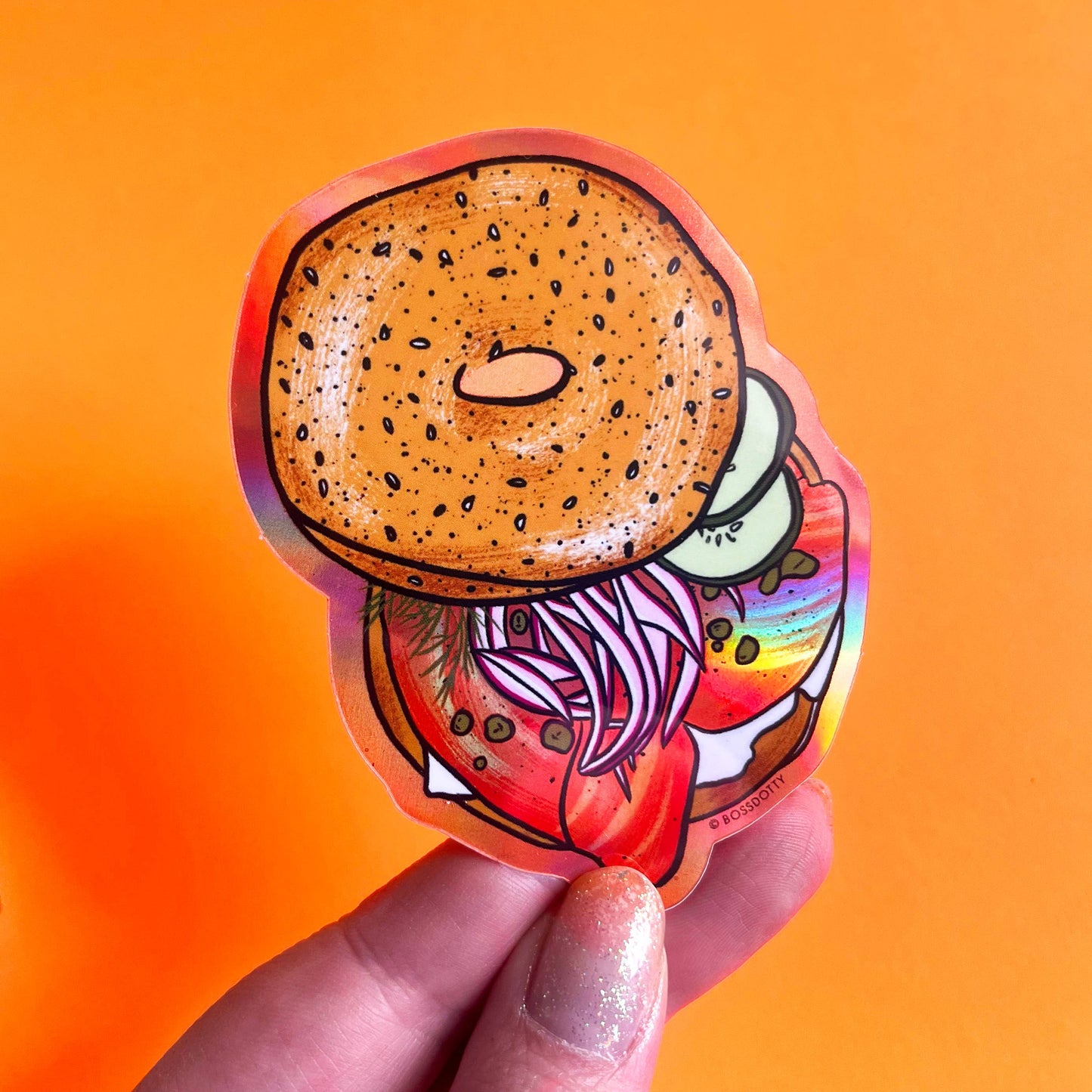 Holographic sticker of a bagel with various toppings held against an orange background