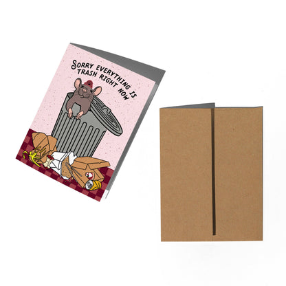 A greeting card and matching kraft envelope on a white background. The card features a pink background with a cartoon mouse sitting on a gray trash can, surrounded by trash, with the text 'Sorry EVERYTHING IS TRASH RIGHT NOW!'.