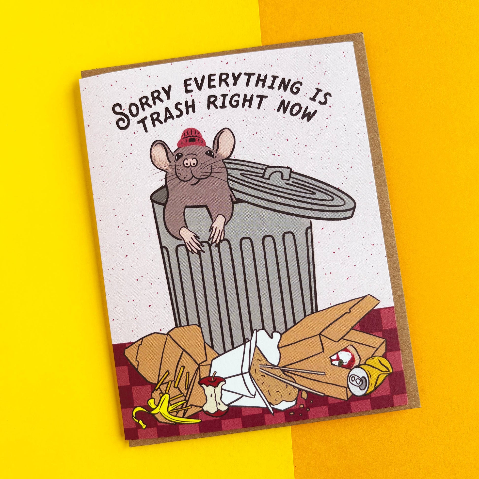 A rectangular novelty card or print with a cartoon mouse in a red firefighter helmet leaning on a trash can, with the text 'SORRY EVERYTHING IS TRASH RIGHT NOW' above. The card is photographed on a bright yellow background.