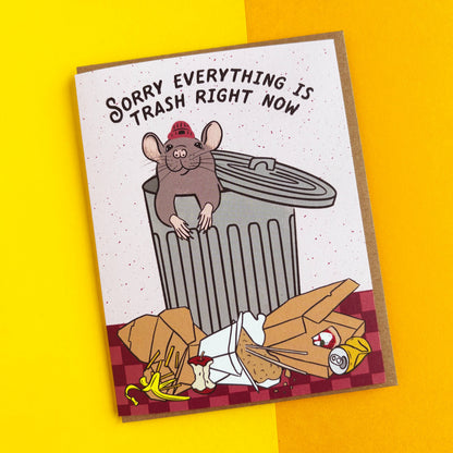 A rectangular novelty card or print with a cartoon mouse in a red firefighter helmet leaning on a trash can, with the text 'SORRY EVERYTHING IS TRASH RIGHT NOW' above. The card is photographed on a bright yellow background.