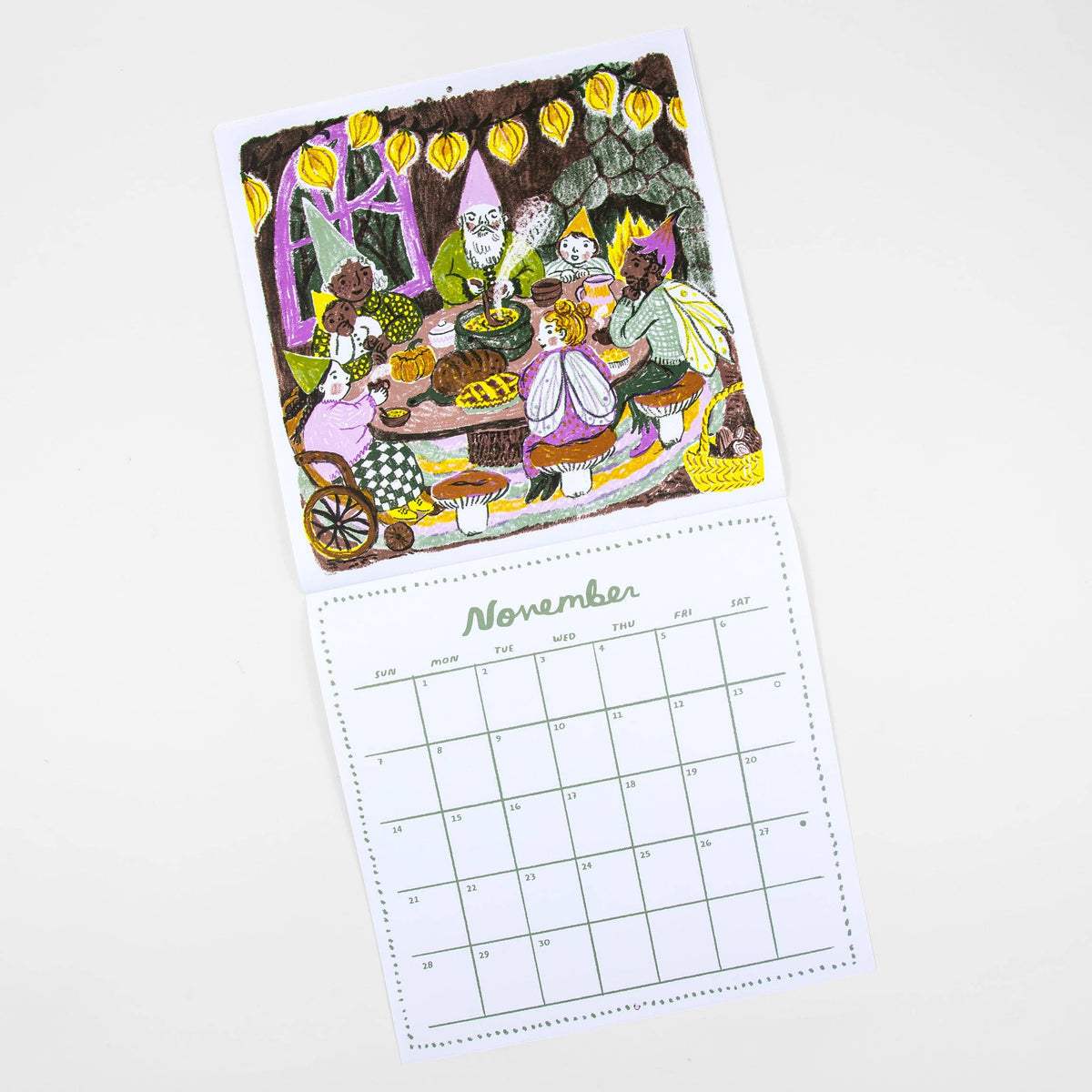 A single-page desk calendar card shown on a white background, featuring a whimsical illustrated scene of garden gnomes gathered around a table with autumn foods, above a printed November calendar grid.