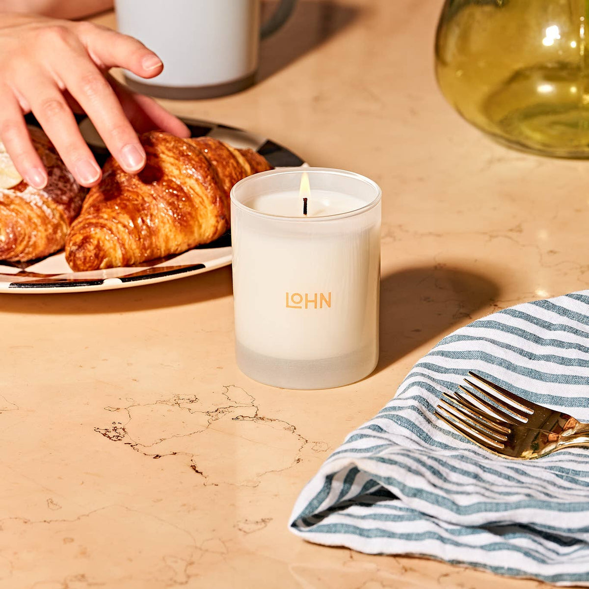 Candle with 'LOHN' branding on a table with croissants and a striped cloth.