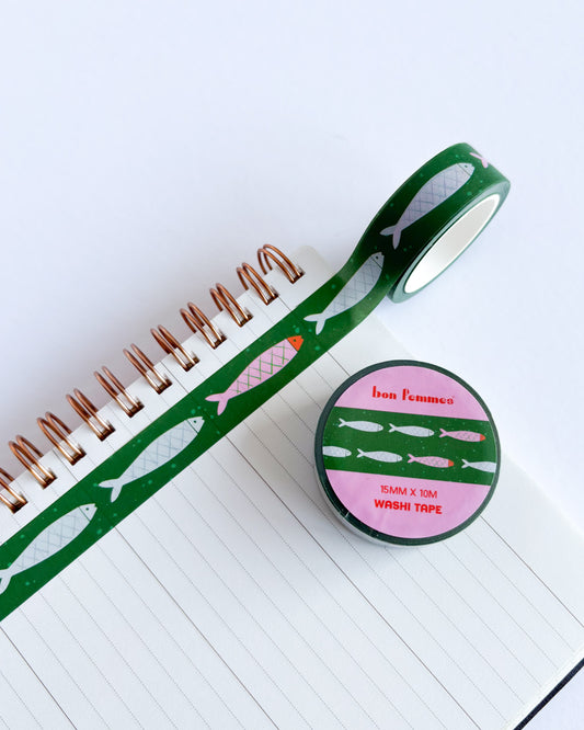 A roll of green washi tape with fish illustrations is unrolled across a lined notebook, with the matching round container lid placed nearby on a white background.