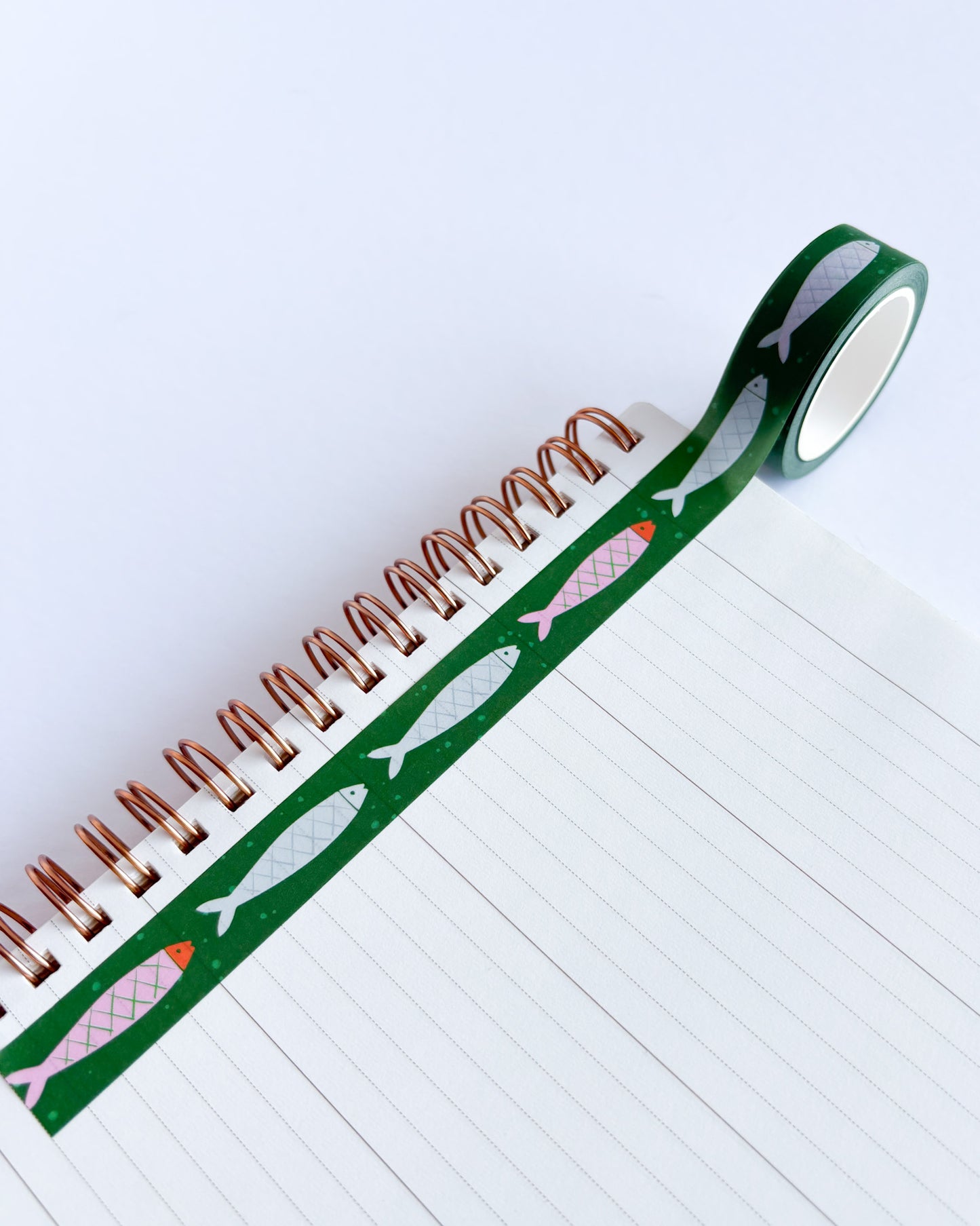 A roll of green washi tape with a pattern of silver, pink, and orange fish, partially unrolled over a white lined spiral notebook on a clean white background.