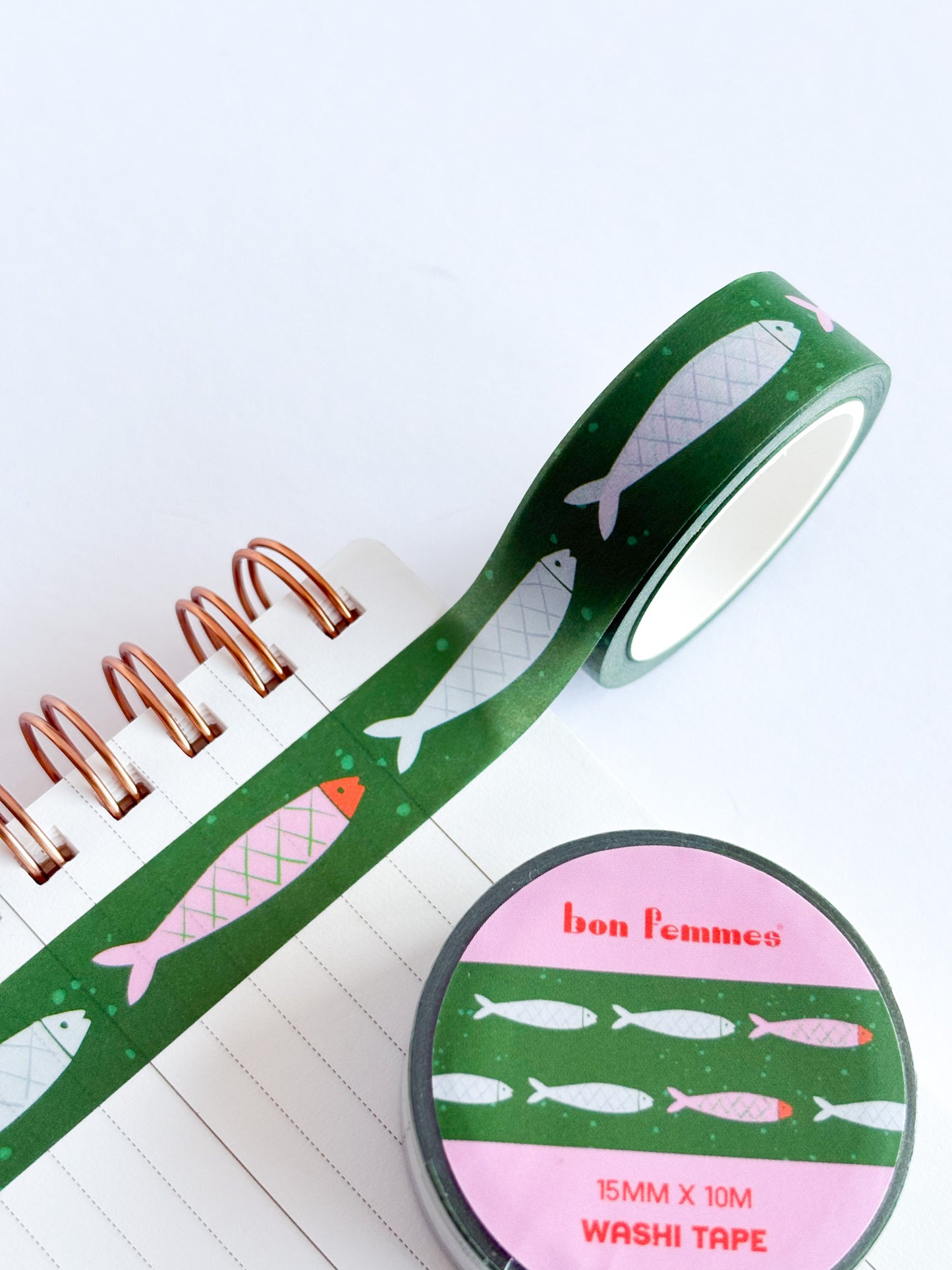 Green washi tape with a fish pattern, shown partially unrolled over a spiral notebook and with its round packaging lid in front. The lid is pink with white fish illustrations and the brand name 'bon femme'.