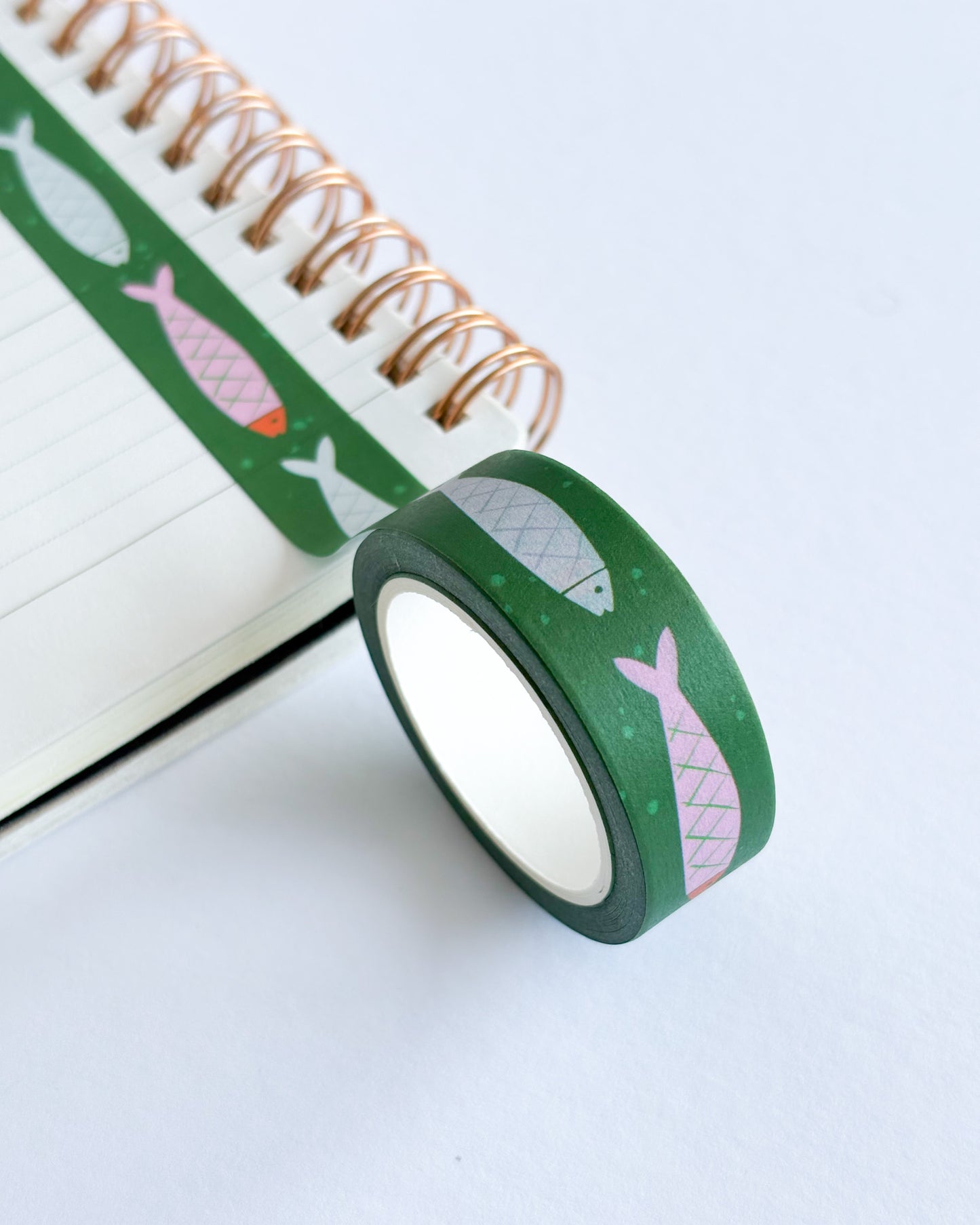 A roll of green washi tape with pink and gray fish patterns, shown next to an open spiral notebook on a clean white background.