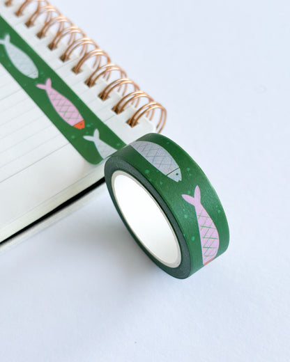 A roll of green washi tape with pink and gray fish patterns, shown next to an open spiral notebook on a clean white background.