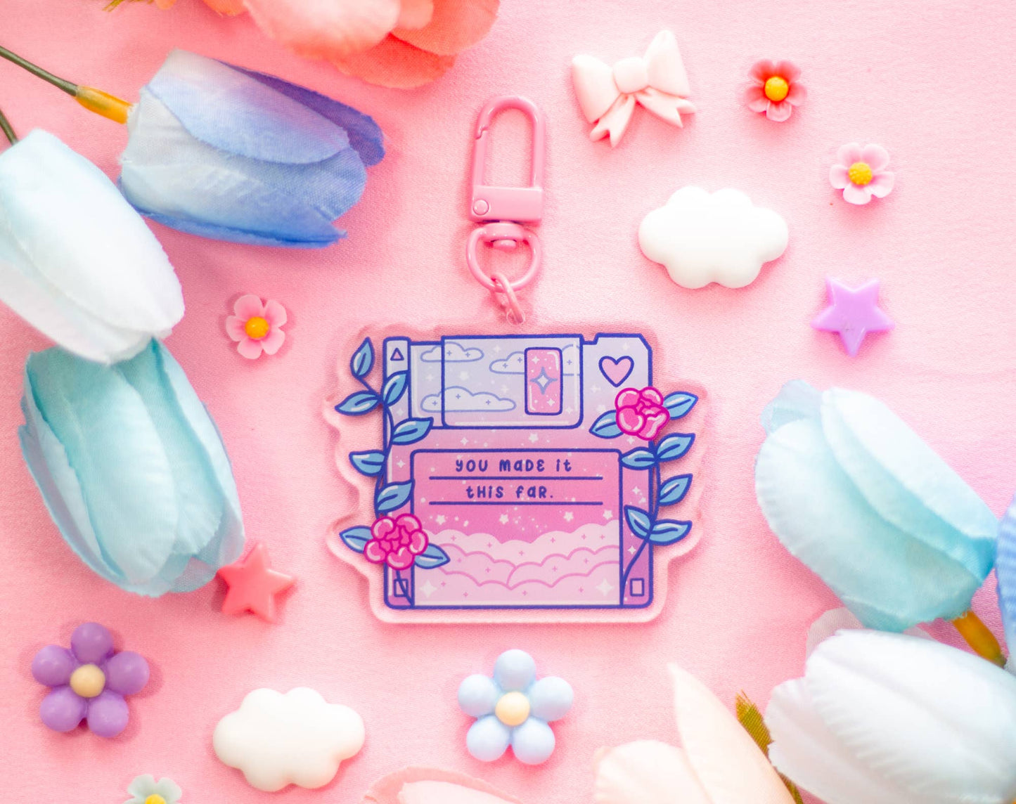 A pastel pink keychain or bag charm shaped like a retro computer floppy disk, decorated with blue leaves, pink flowers, and the phrase 'YOU MADE IT THIS FAR.' It is photographed on a pink background with decorative flowers, clouds, bows, and stars around it.